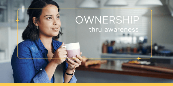 A young woman sits in a kitchen, hands wrapped around a coffee cup, gazing thoughtfully into the distance as she pauses to become aware of her emotions—practicing a simple yet powerful technique for emotional regulation