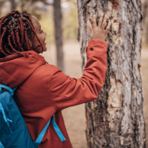 Young adult in red jacket touches tree during Flagstaff Forest Bathing, surrounded by tall pines.